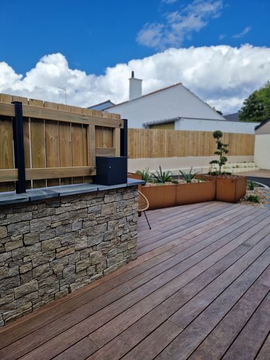 Terrasse en bois avec jardinières surélevées, muret en pierre et clôture sous un ciel bleu.