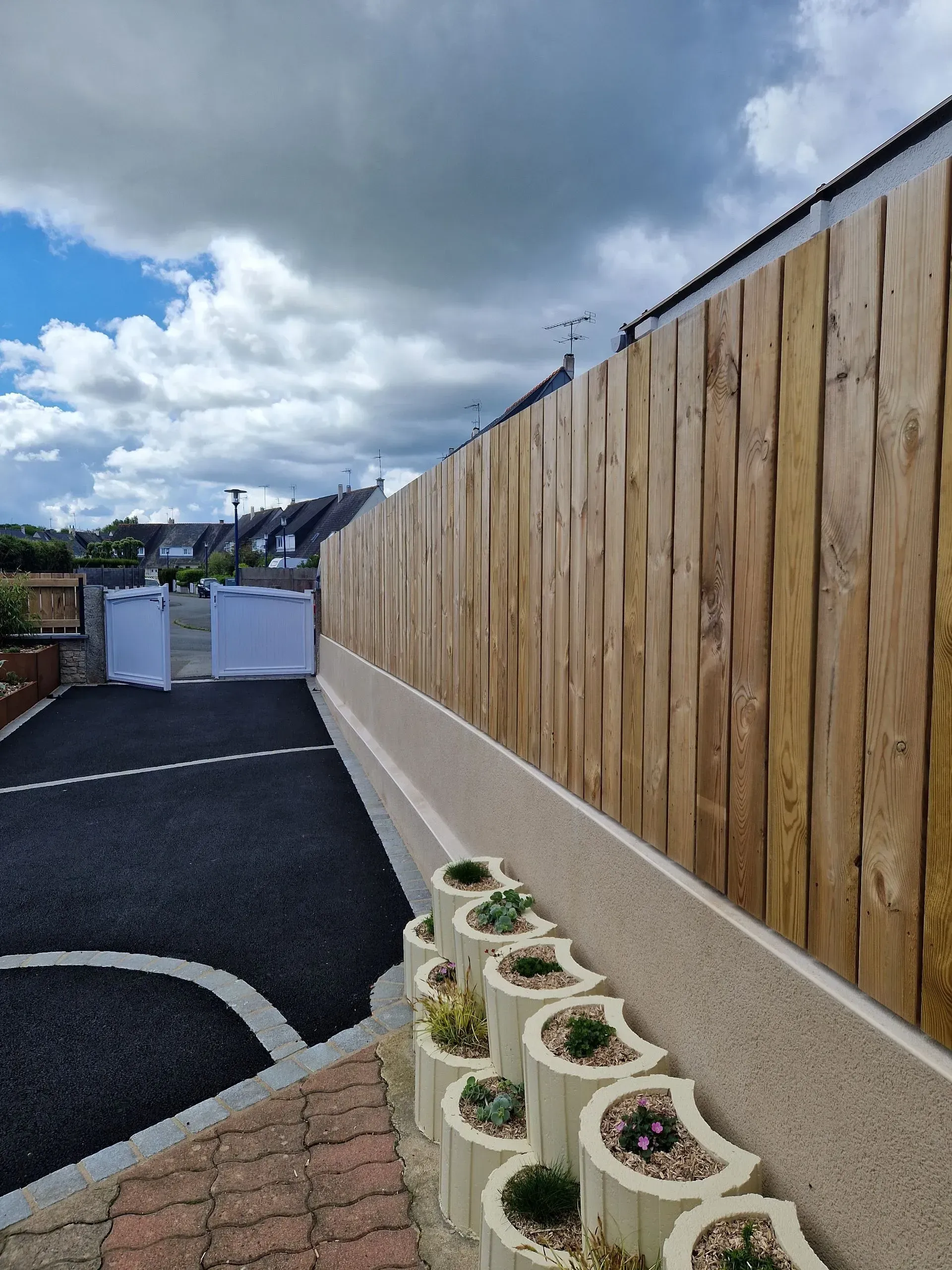 Allée longeant une haute clôture en bois, avec des pots de fleurs et un ciel nuageux au-dessus.