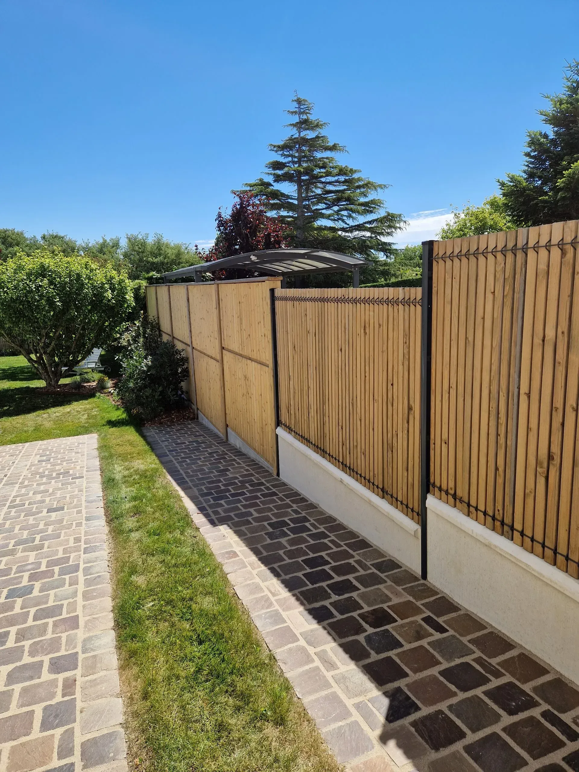 Clôture en bois longeant un chemin de jardin ensoleillé, bordé d'une pelouse et de pavés.