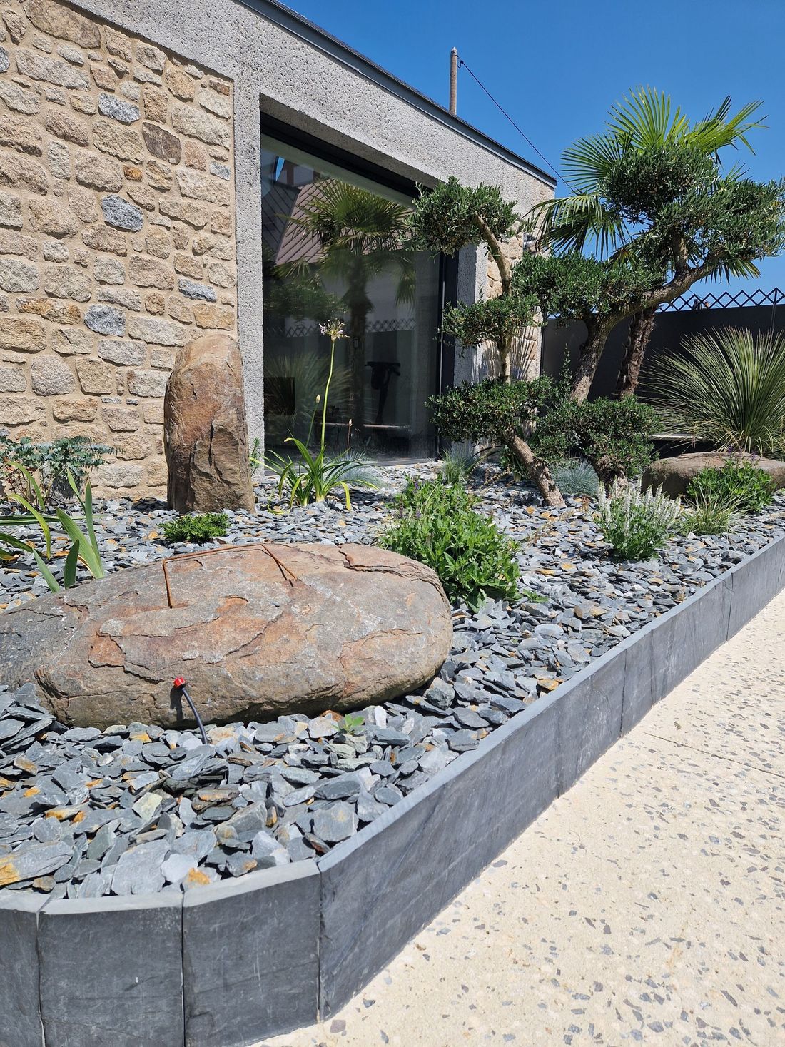 Entrée d'un bâtiment en pierre avec jardin de gravier, gros rocher et petit arbre au bord d'un chemin pavé.