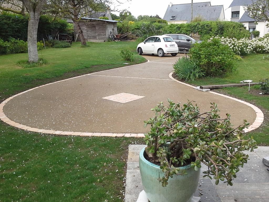 Une allée circulaire en gravier dans un jardin, avec une voiture blanche garée près d'arbustes et une plante en pot au premier plan.