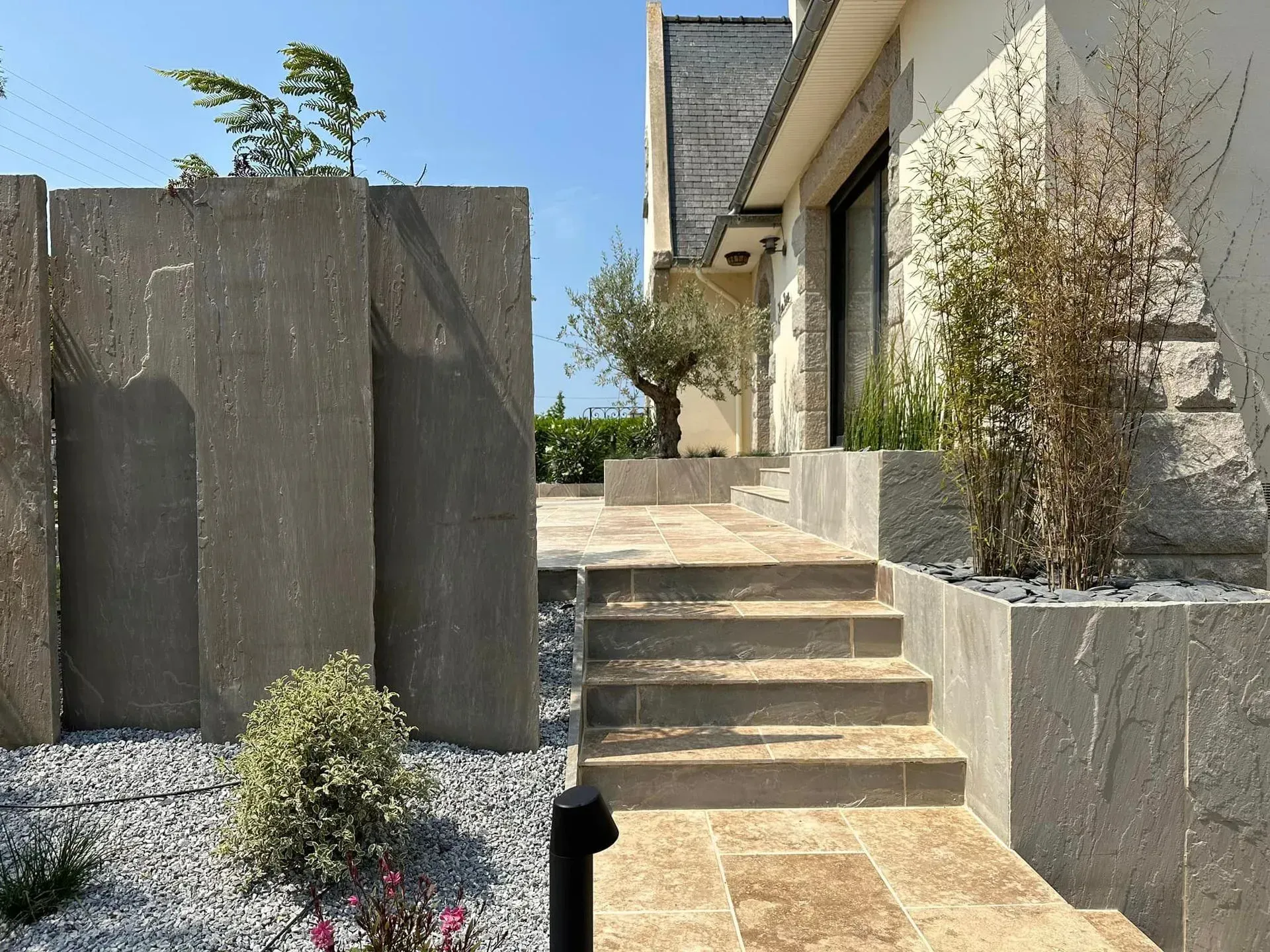Des marches en béton mènent à l'entrée d'une maison moderne avec des murs en pierre et un aménagement paysager.