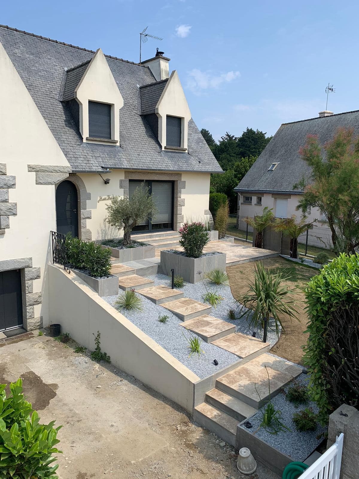 Maison en pierre au toit gris, avec un petit patio paysager à l'avant, agrémenté de plantes en pot et d'un escalier.