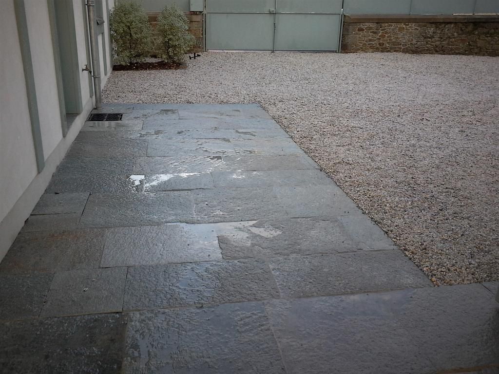 Terrasse en pierre jouxtant une cour en gravier et un mur d'enceinte, avec une zone humide près du centre.