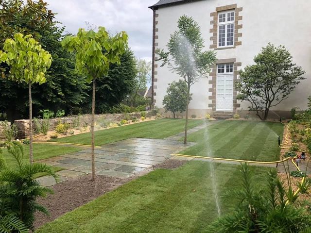 Jardin avec arroseurs automatiques arrosant une pelouse jouxtant une maison blanche entourée d'arbres et bordée de pierres.