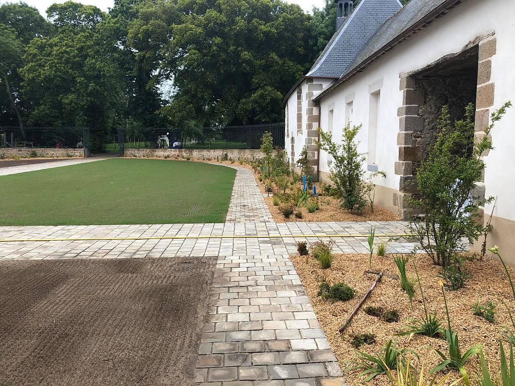 Terrasse en briques jouxtant une maison blanche, avec parterres de fleurs aménagés, pelouse et arbres en arrière-plan.