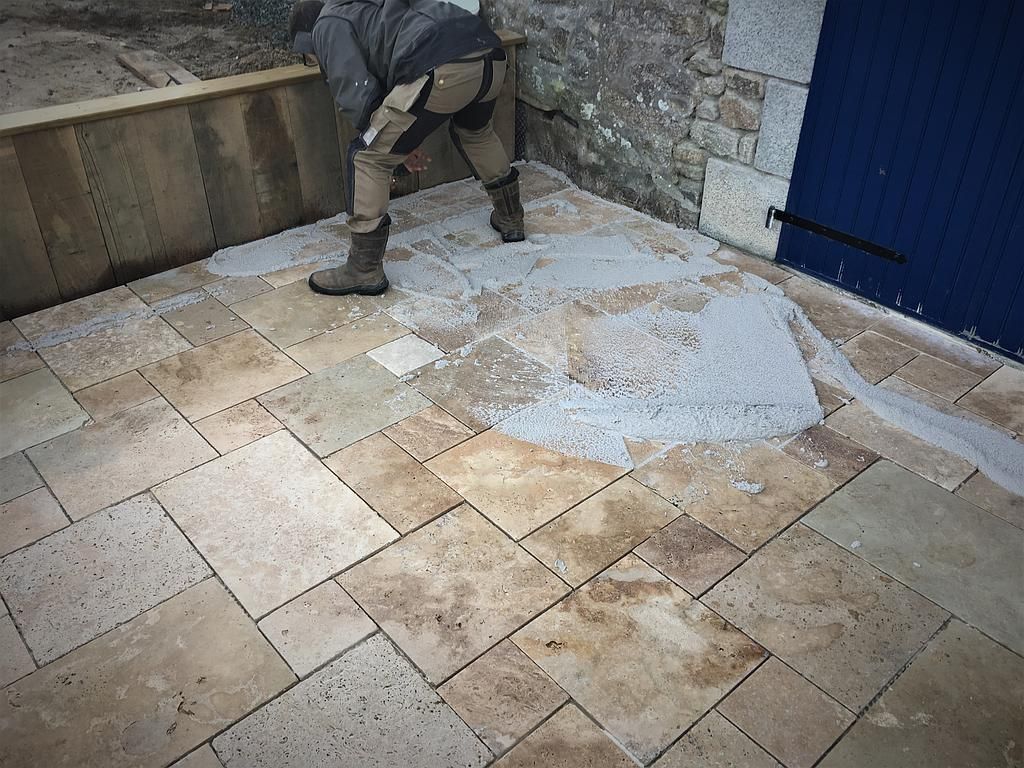 Un ouvrier pose du carrelage sur une terrasse en pierre le long d'un mur, avec une porte bleue et une plaque de béton visibles.