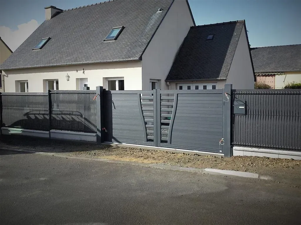 Maison grise derrière une clôture et un portail métalliques, au bord d'une rue pavée.