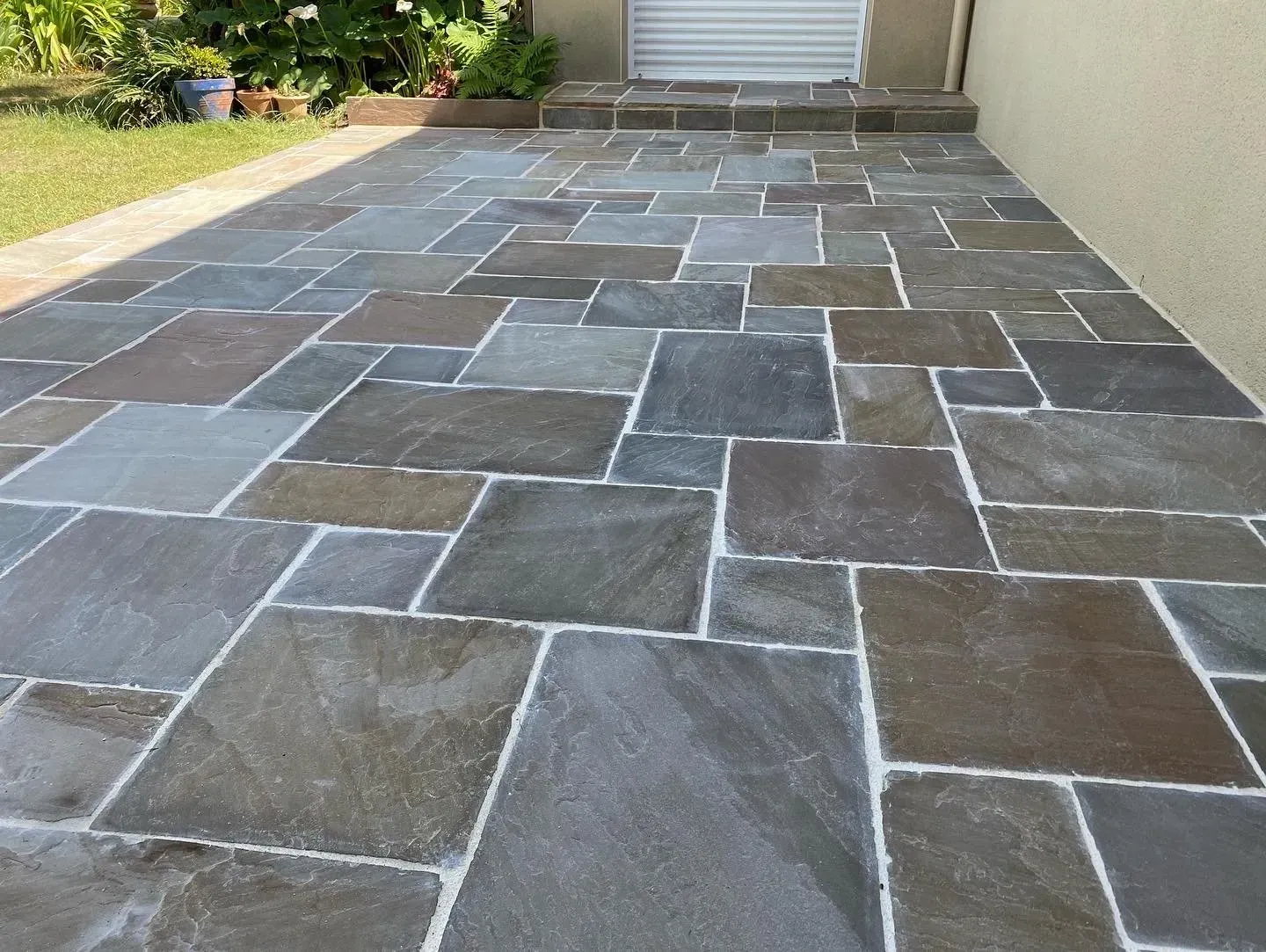 Terrasse arrière pavée de pierres menant à un garage, avec de grands carreaux gris et beiges.