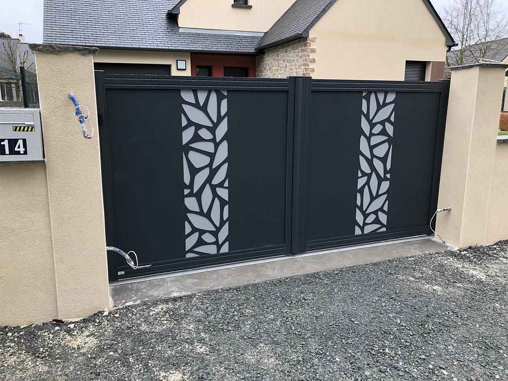 Portail double en métal noir avec panneaux découpés en forme de feuilles devant une maison beige et une allée en gravier.