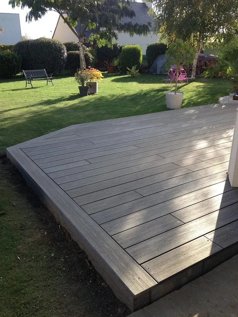 Terrasse en bois dans un jardin ensoleillé avec pelouse verte, bancs, arbustes et fleurs en pot.