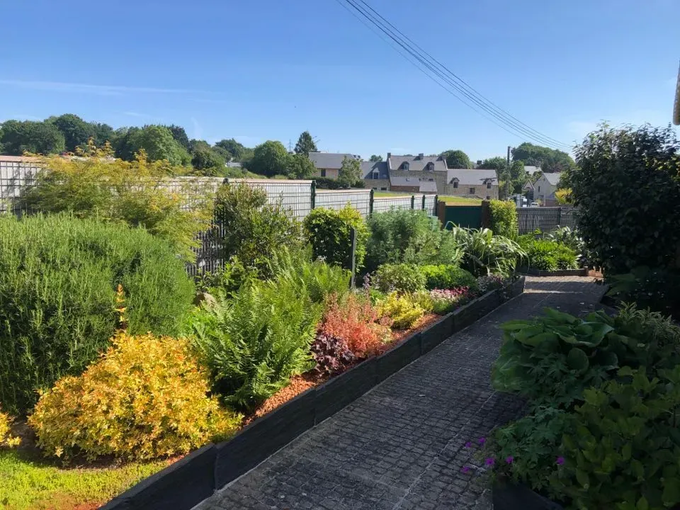 Allée ensoleillée dans un jardin bordée de parterres de fleurs colorés et de maisons en arrière-plan.