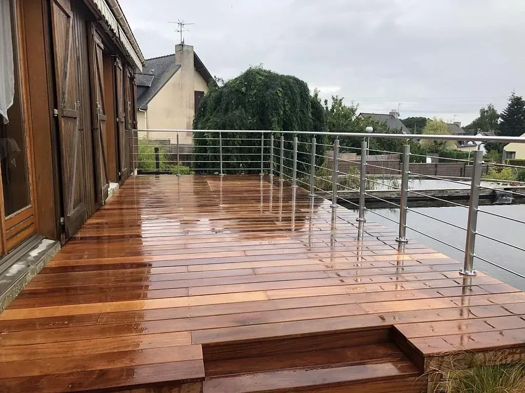 Terrasse en bois sur le toit, finition brillante et mouillée, avec rambarde en métal donnant sur les maisons et les arbres.