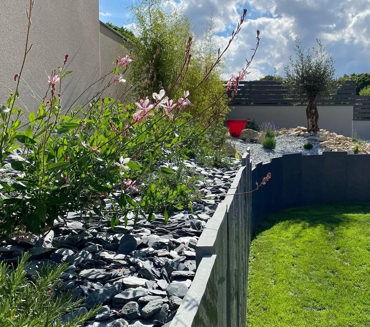 Jardin paysager avec bordure de pierres grises, fleurs roses, pelouse verte et chaise rouge sous un ciel bleu.