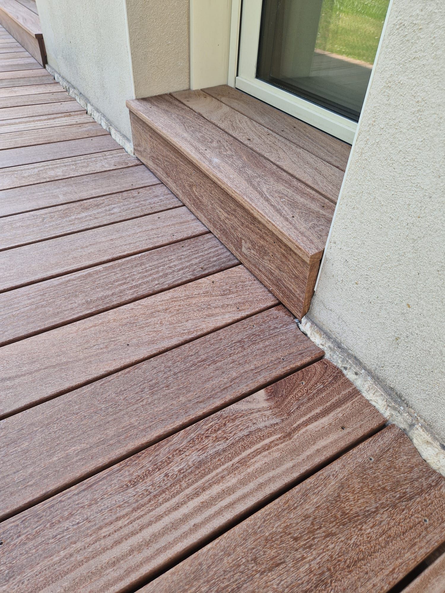 Des planches de terrasse en bois mènent à une petite marche et à une porte coulissante en verre, le long d'un mur en stuc.