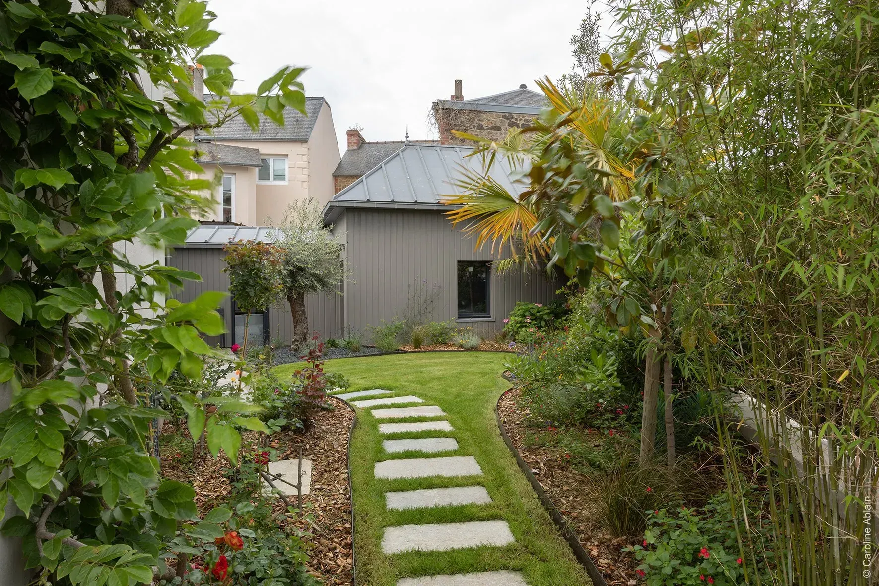 Allée de jardin avec des dalles menant à une petite maison, entourée de verdure.
