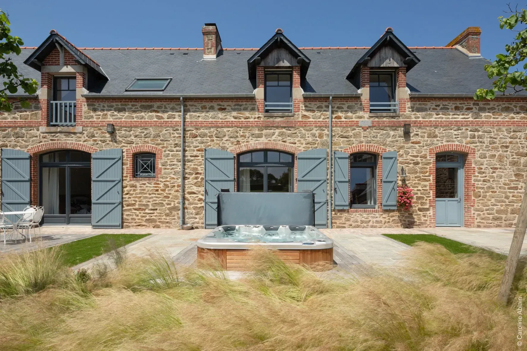 Maison en pierre avec toit noir, lucarnes et terrasse donnant sur un jardin gazonné par une journée ensoleillée.
