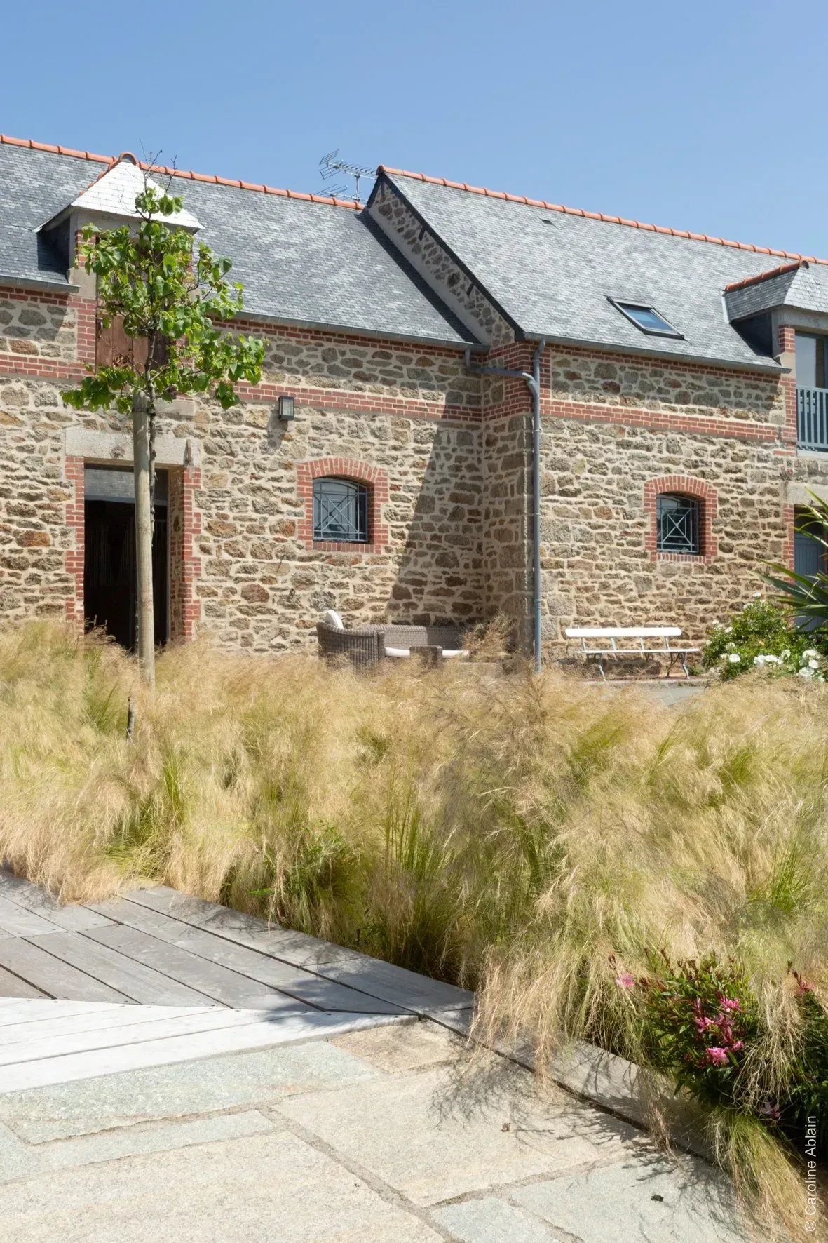 Bâtiment en pierre au toit gris, aux petites fenêtres, et à l'avant, de hautes herbes sèches sous un ciel dégagé.