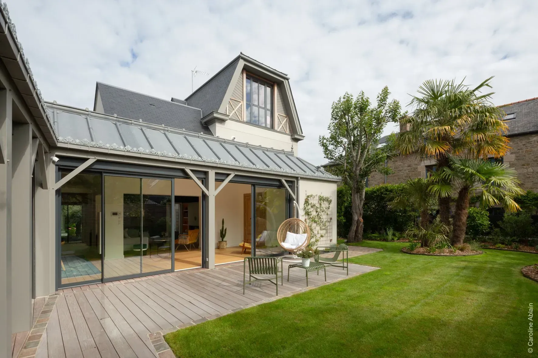 Maison moderne avec de grandes portes vitrées s'ouvrant sur une terrasse en bois et un jardin arrière verdoyant.