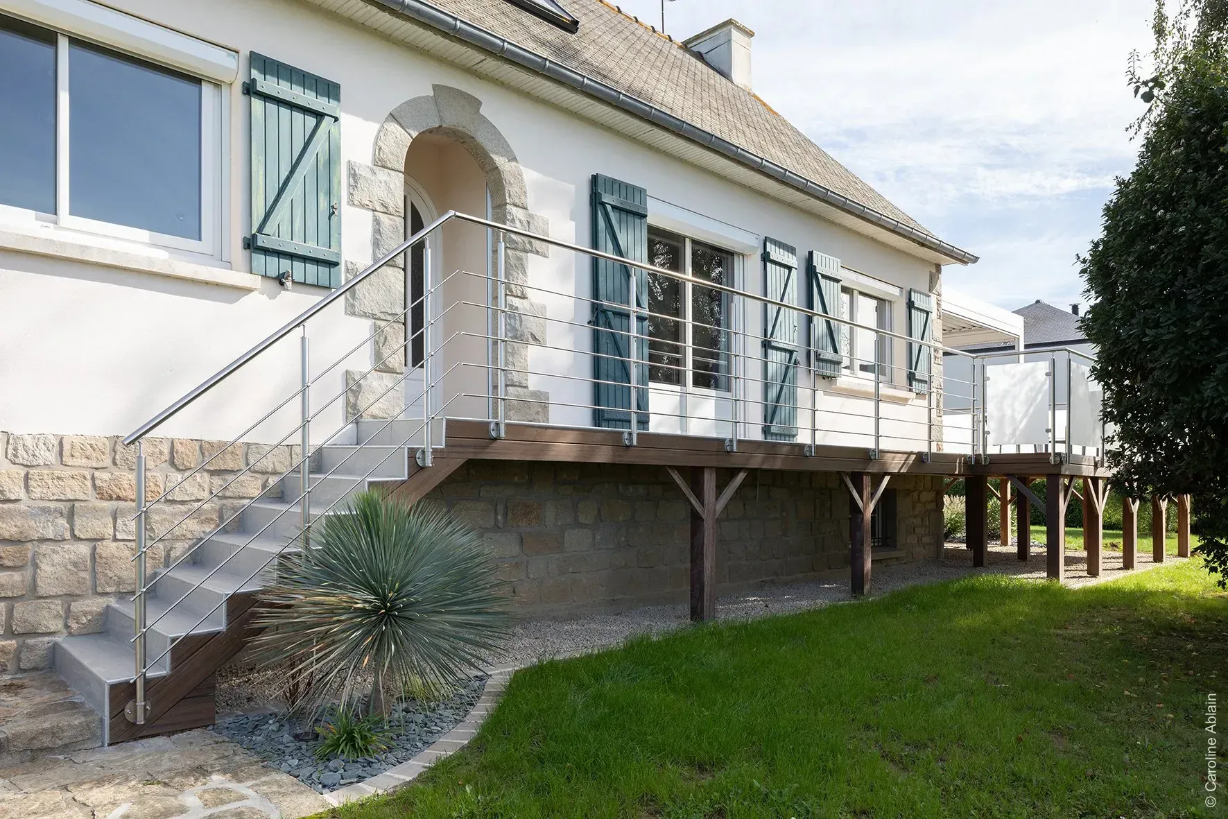 Maison surélevée en pierre et stuc avec volets bleus, escalier extérieur et porche couvert donnant sur une cour gazonnée.