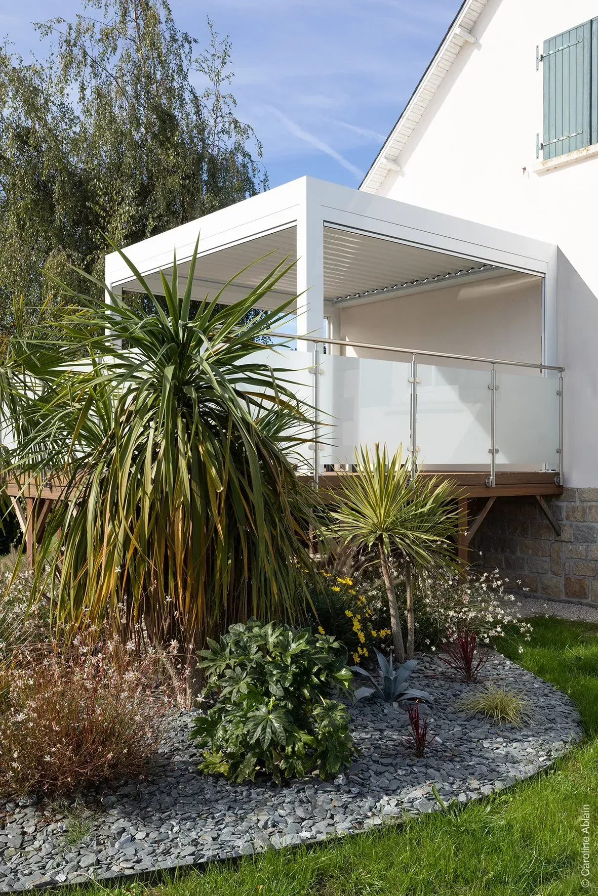 Auvent blanc au-dessus d'un balcon jouxtant une maison, avec des arbustes de jardin luxuriants et du gravier en dessous.