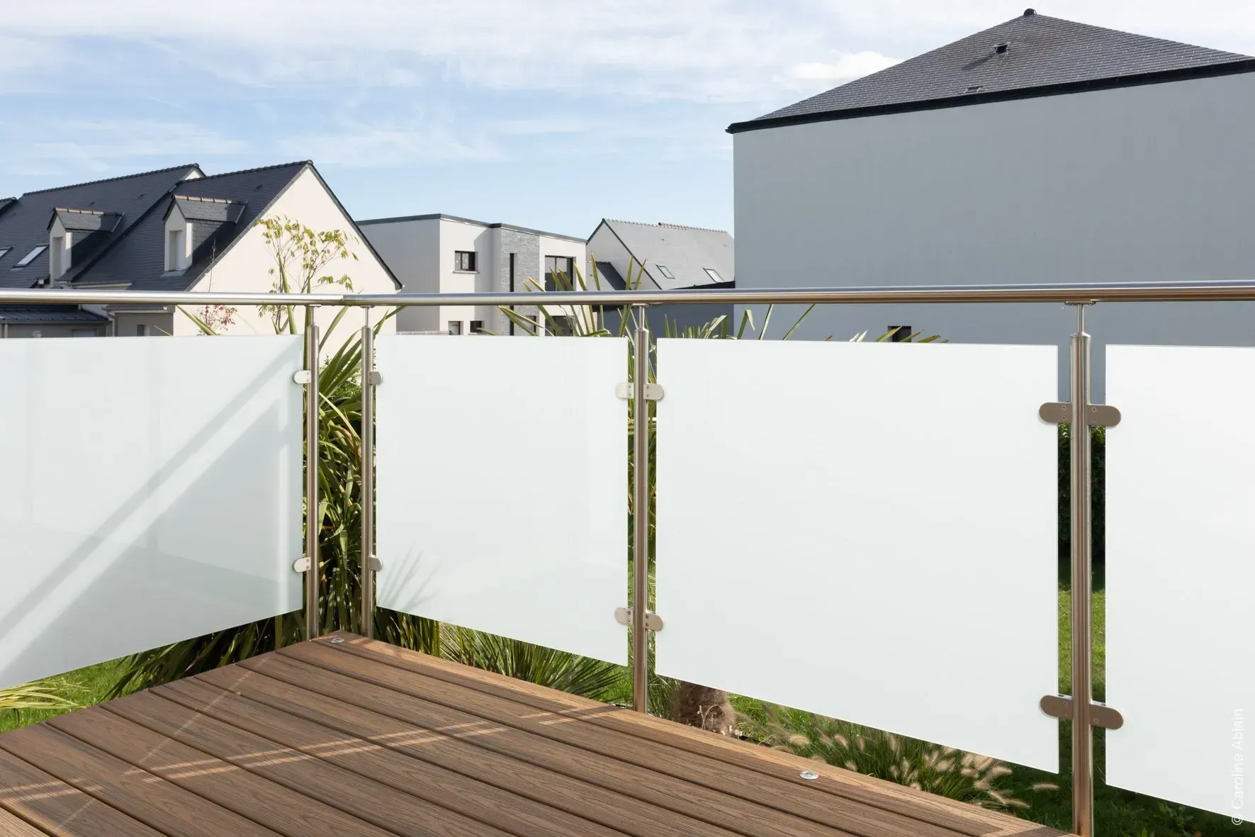 Balcon avec balustrade en verre et panneaux dépolis sur une terrasse en bois, donnant sur les maisons voisines.
