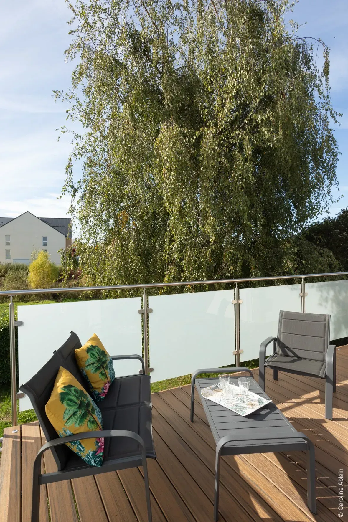 Terrasse aménagée avec coussins et table le long d'une balustrade en verre, donnant sur un grand arbre feuillu et la maison voisine.
