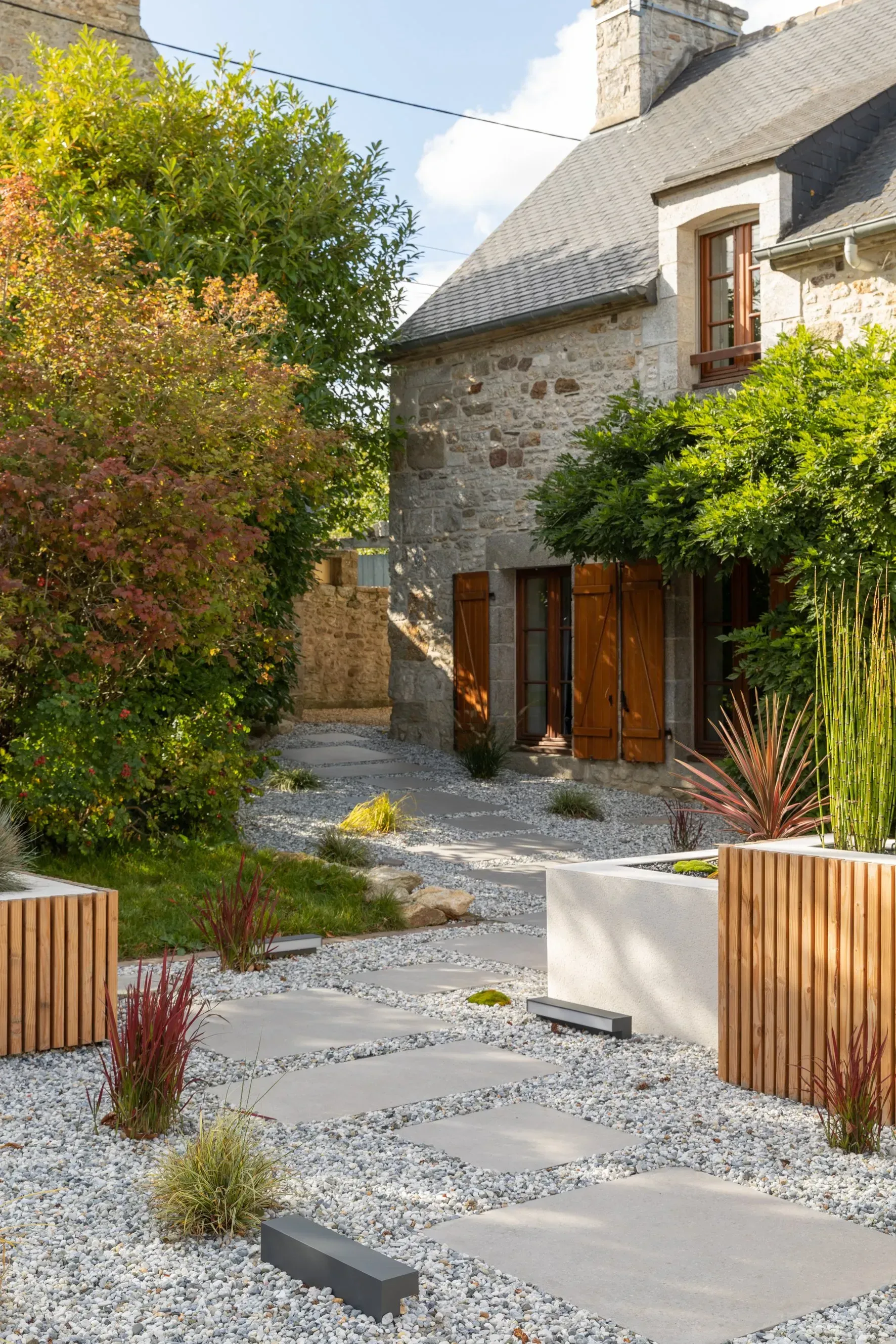 Cour intérieure en pierre avec allée de gravier, plantes en pot, jardinières en bois et façade de maison rustique.