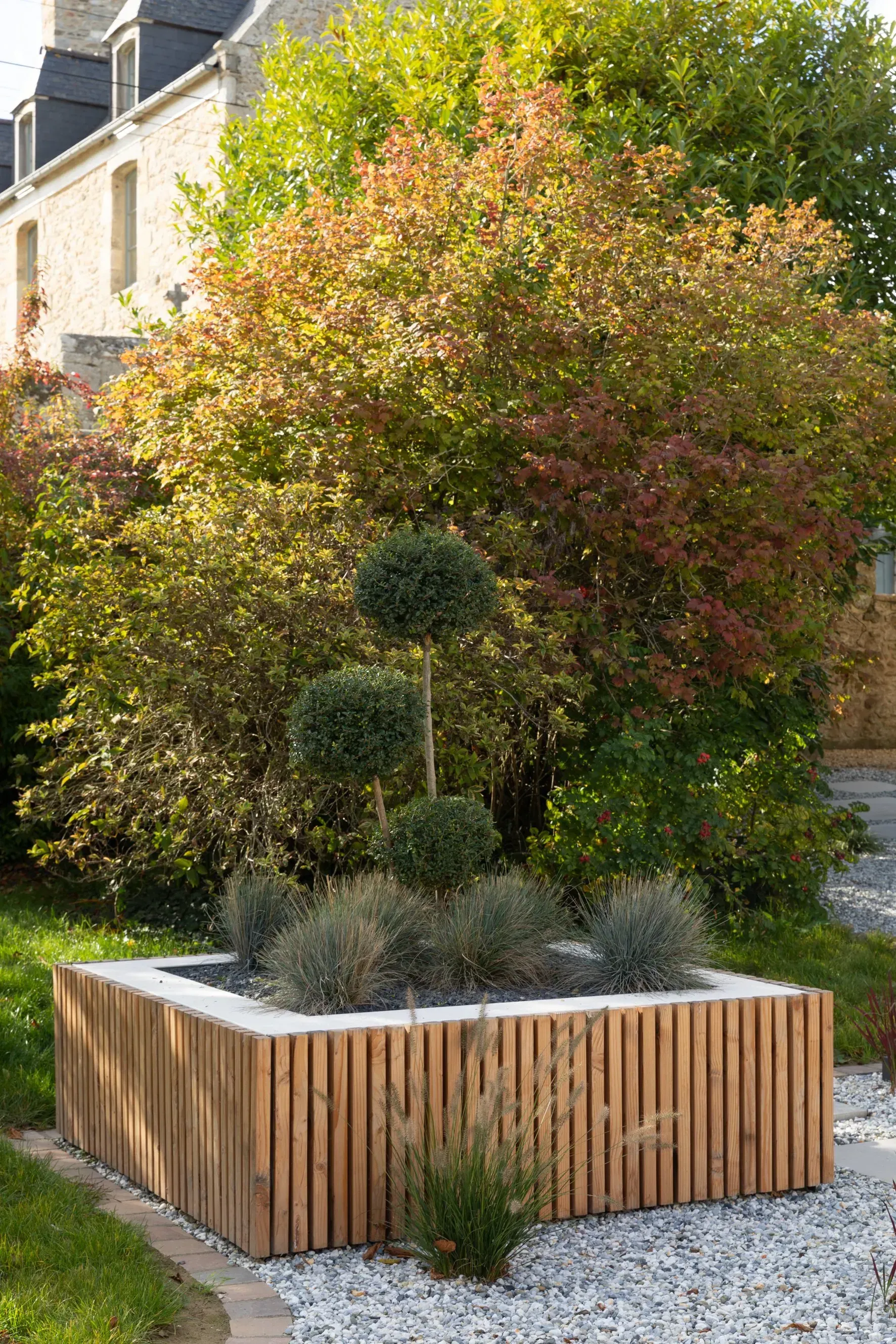 Jardinière moderne en bois avec arbustes ornementaux et petit arbre dans une cour gravillonnée.