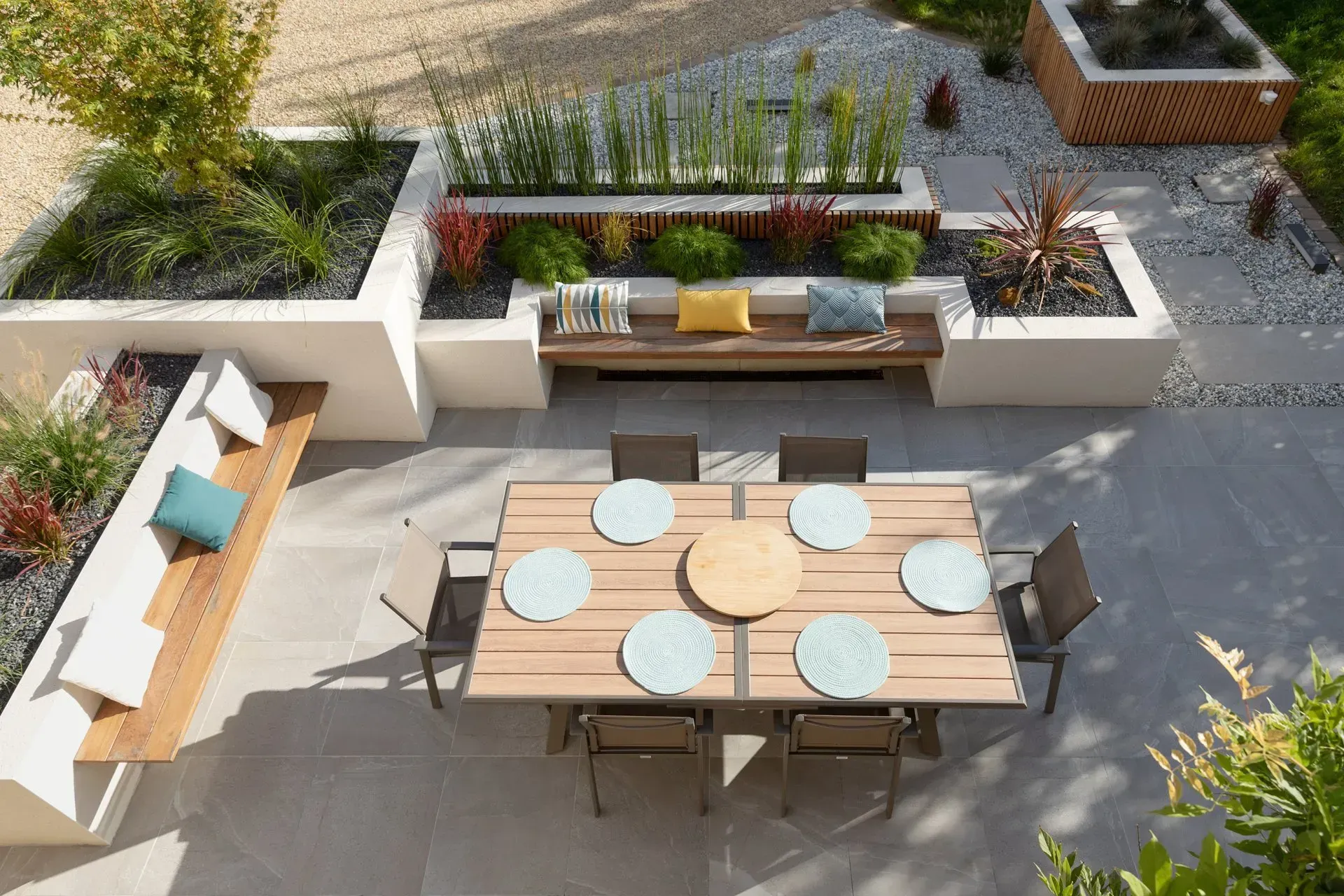 Terrasse arrière ensoleillée avec de grandes dalles carrées en pierre et des joints foncés, bordée de gazon et d'arbustes.