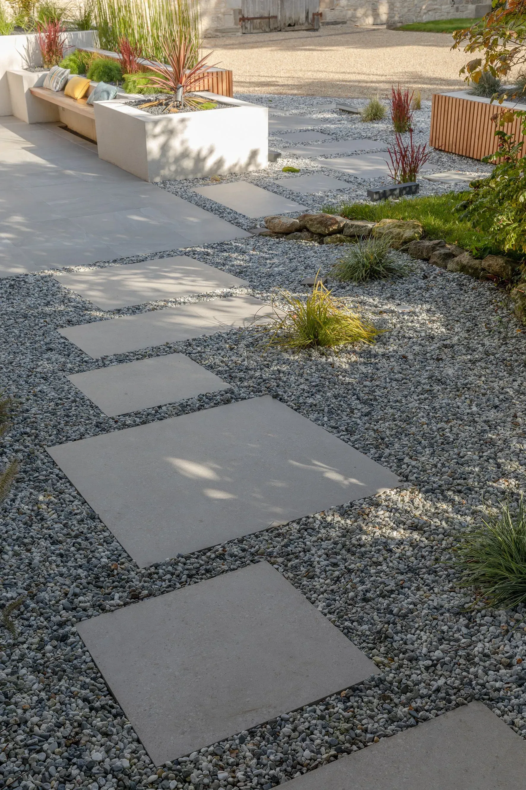 Chemin de pierres traversant un jardin de gravier avec des jardinières surélevées et de la verdure.