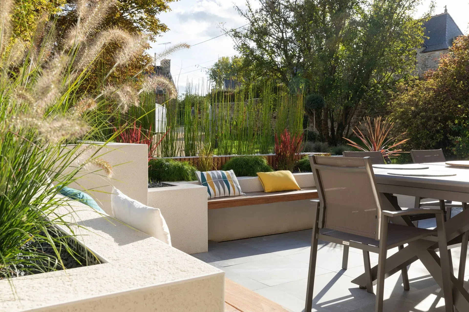 Terrasse ensoleillée avec sièges beiges, table à manger et plantes de jardin verdoyantes.