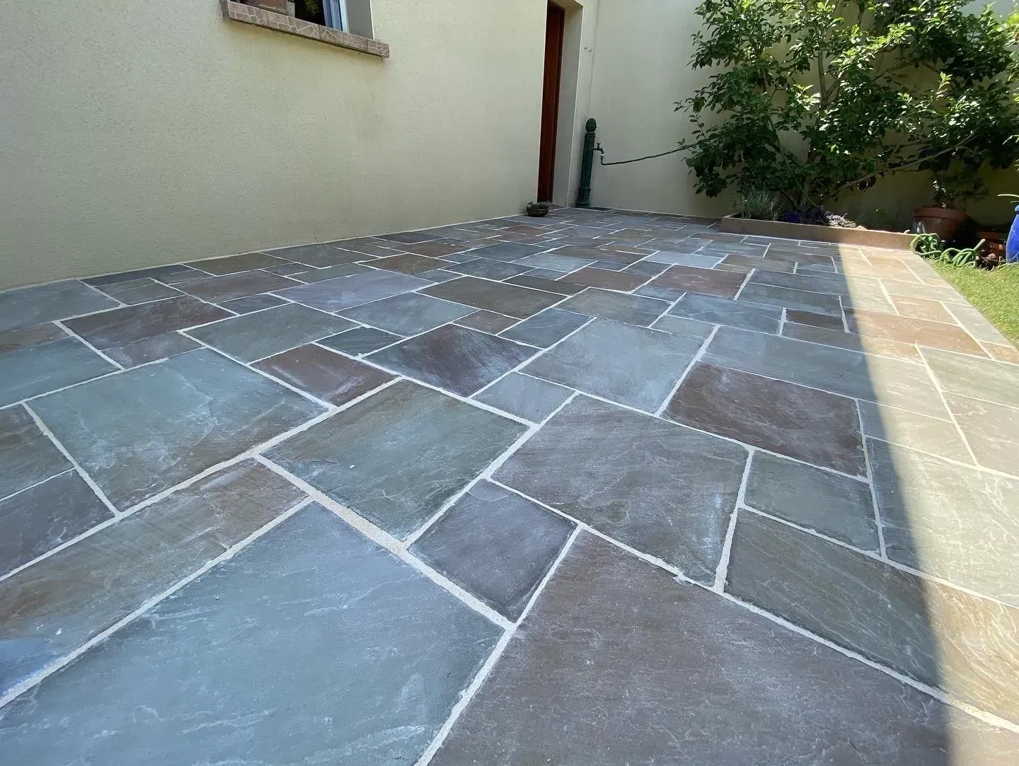 Terrasse carrelée en pierre le long d'un mur de maison, avec un jardin et des buissons en arrière-plan.