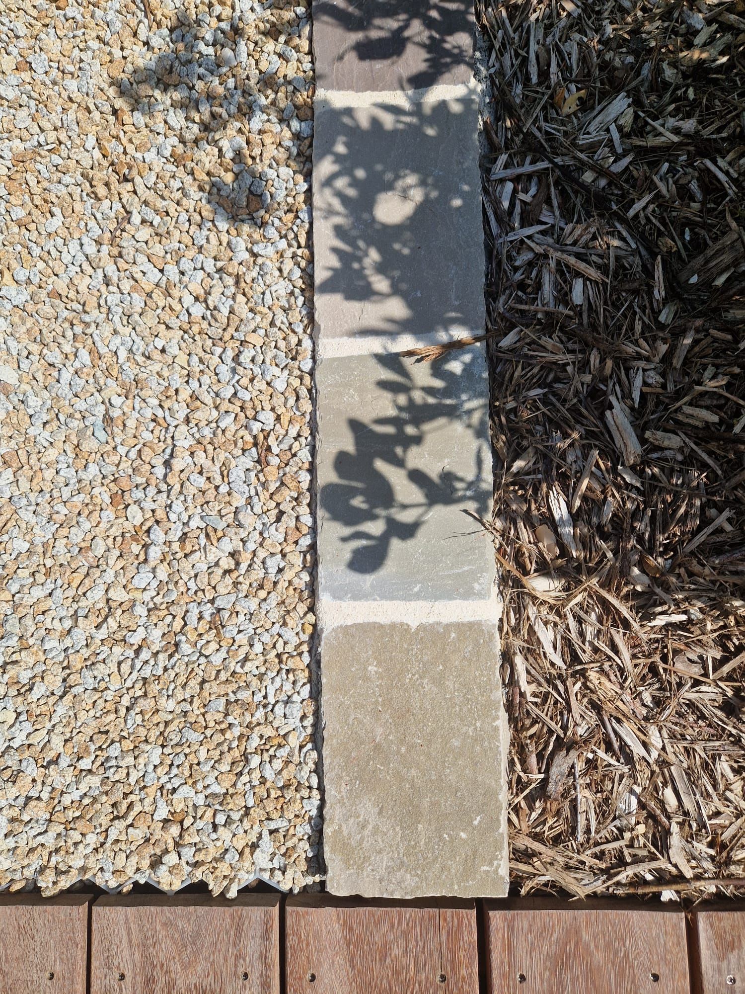 Bordure de jardin en gravier, pavés et paillis d'écorce, avec des ombres de feuilles sur l'allée.