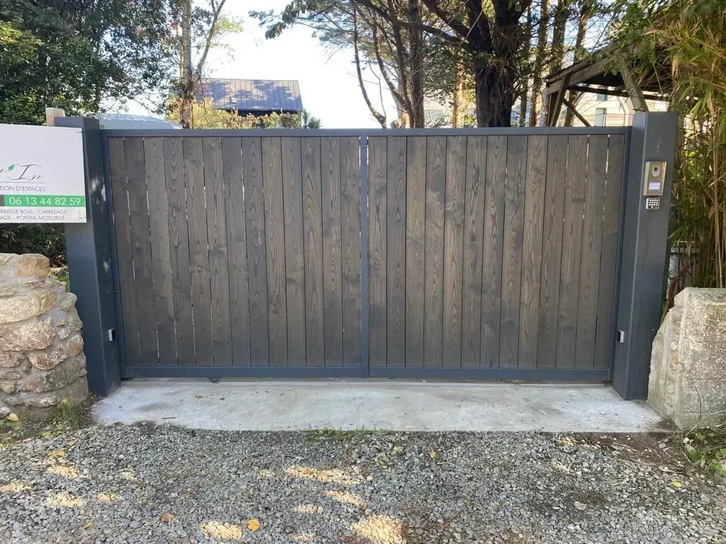 Portail d'allée en bois sombre fermé, avec piliers en pierre et dalle en béton devant une maison.