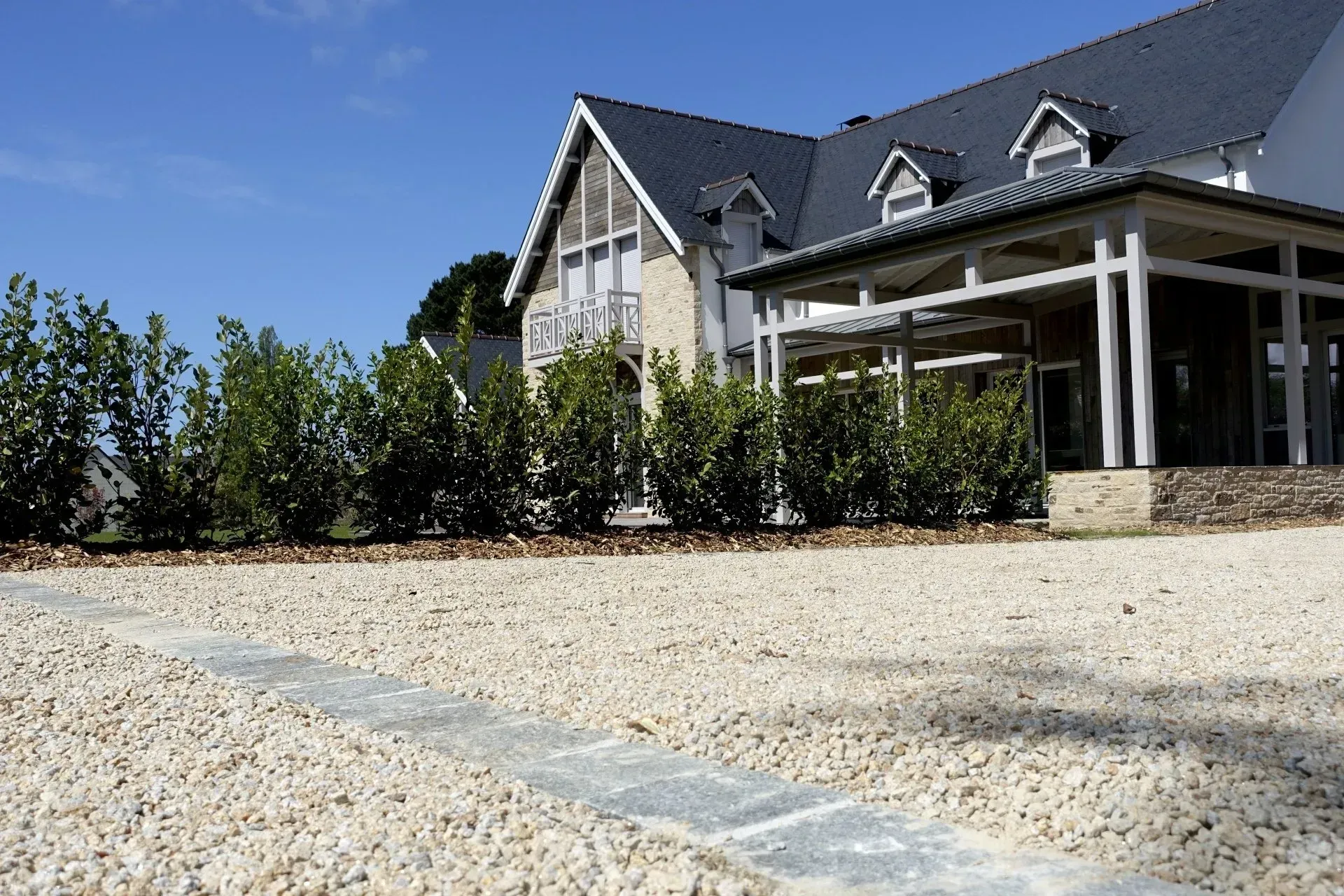 Grande maison avec toit sombre et porche couvert, située à côté d'une allée de gravier et de buissons paysagers.