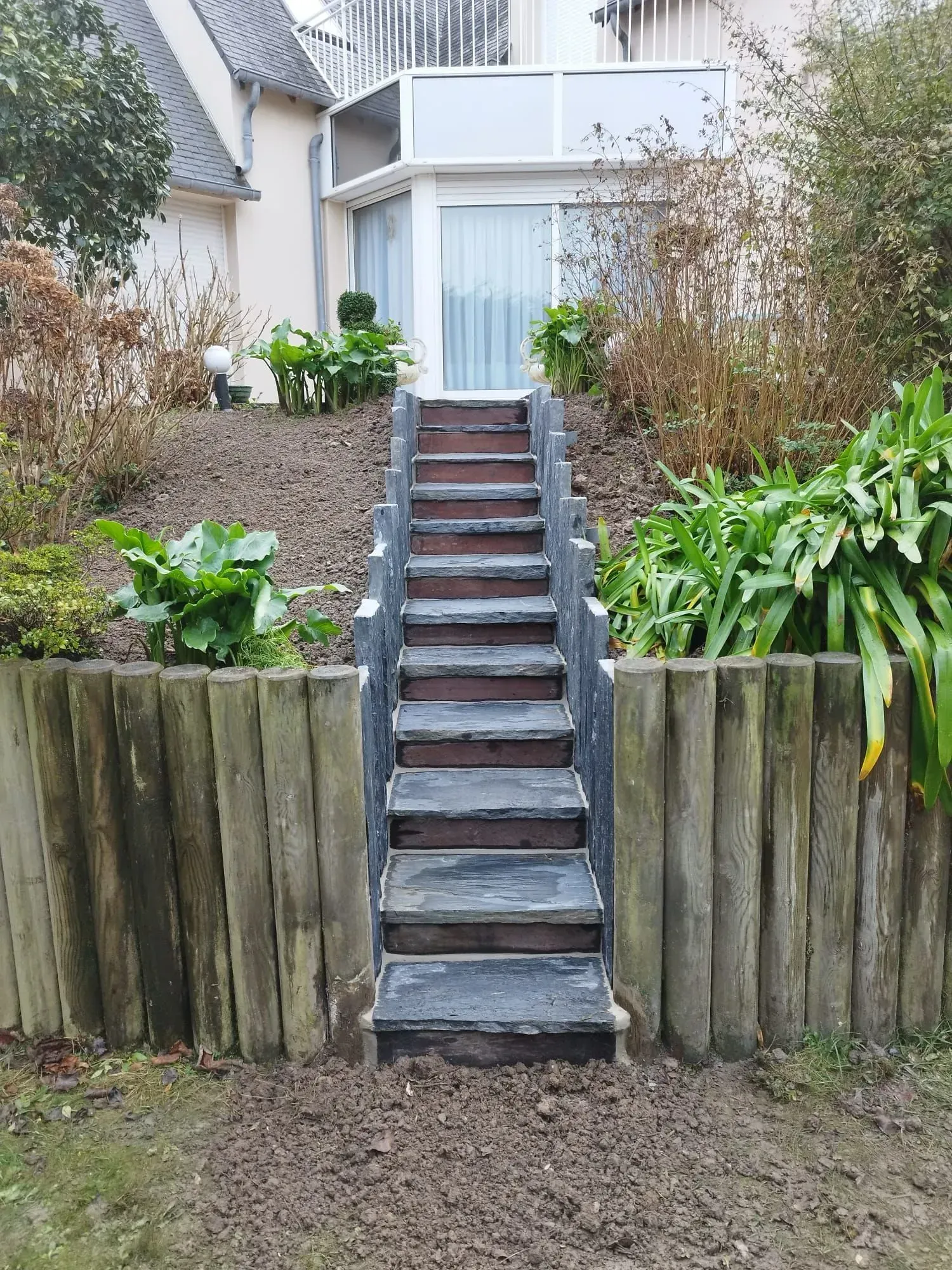 Cour avant avec des marches en bois montant une pente de jardin jusqu'à l'entrée de la maison, bordée de murs de soutènement en bois patiné.