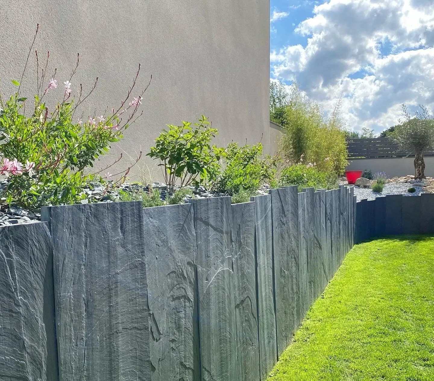 Haute clôture sombre bordée de verdure, longeant une pelouse ensoleillée à l'arrière d'un jardin, près d'un mur beige sous un ciel partiellement nuageux.