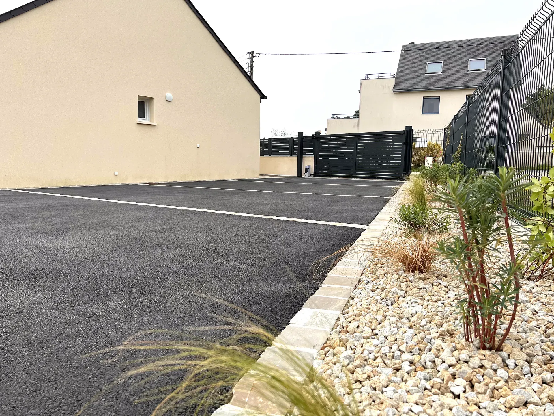 Allée de gravier longeant des maisons beiges avec un portail noir et une bordure paysagée en pierres.