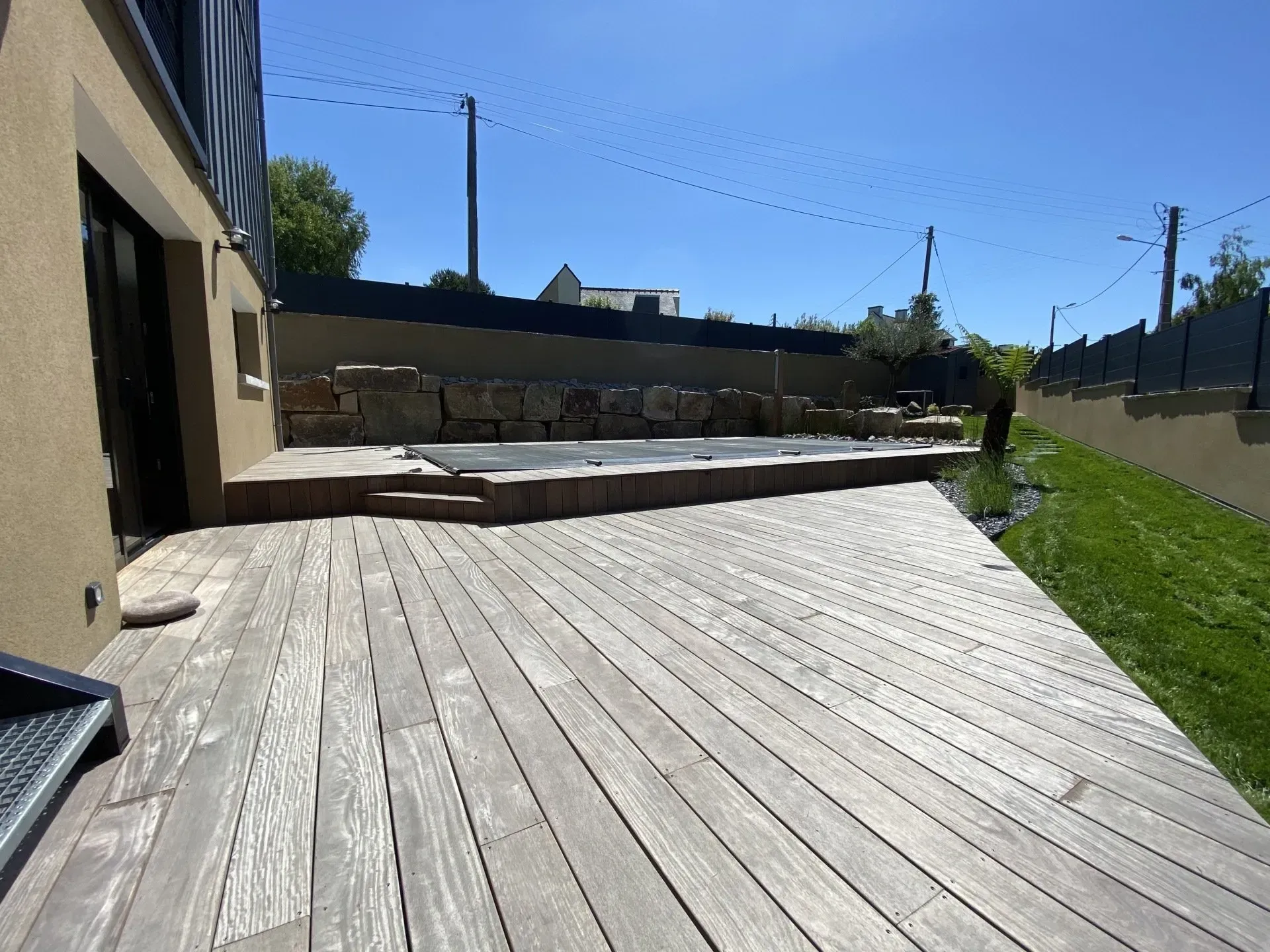 Terrasse en bois ensoleillée jouxtant un bâtiment, avec de l'herbe, un muret et le ciel bleu au-delà.
