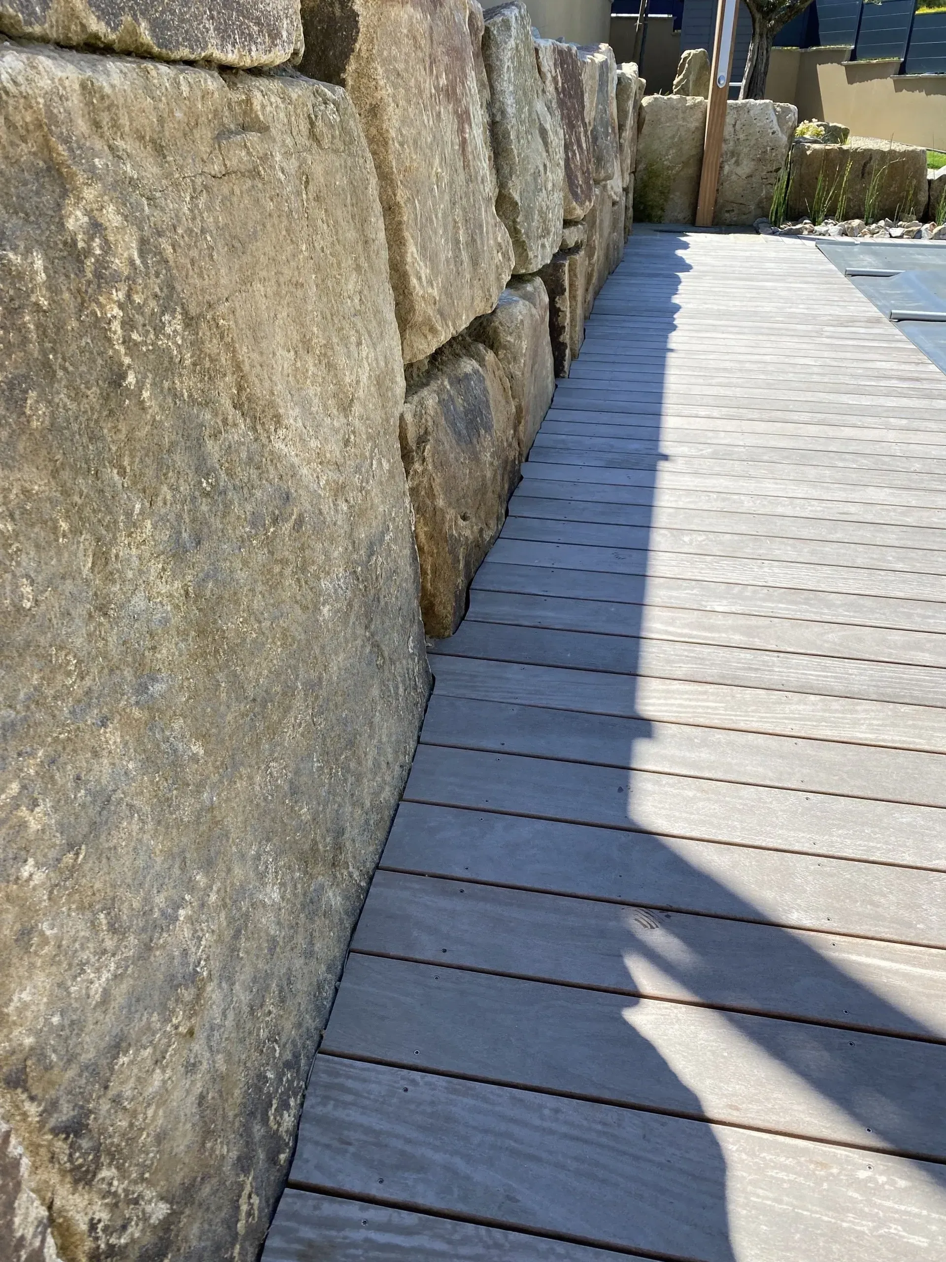 Mur de pierre jouxtant une passerelle en bois ensoleillée aux ombres profondes.