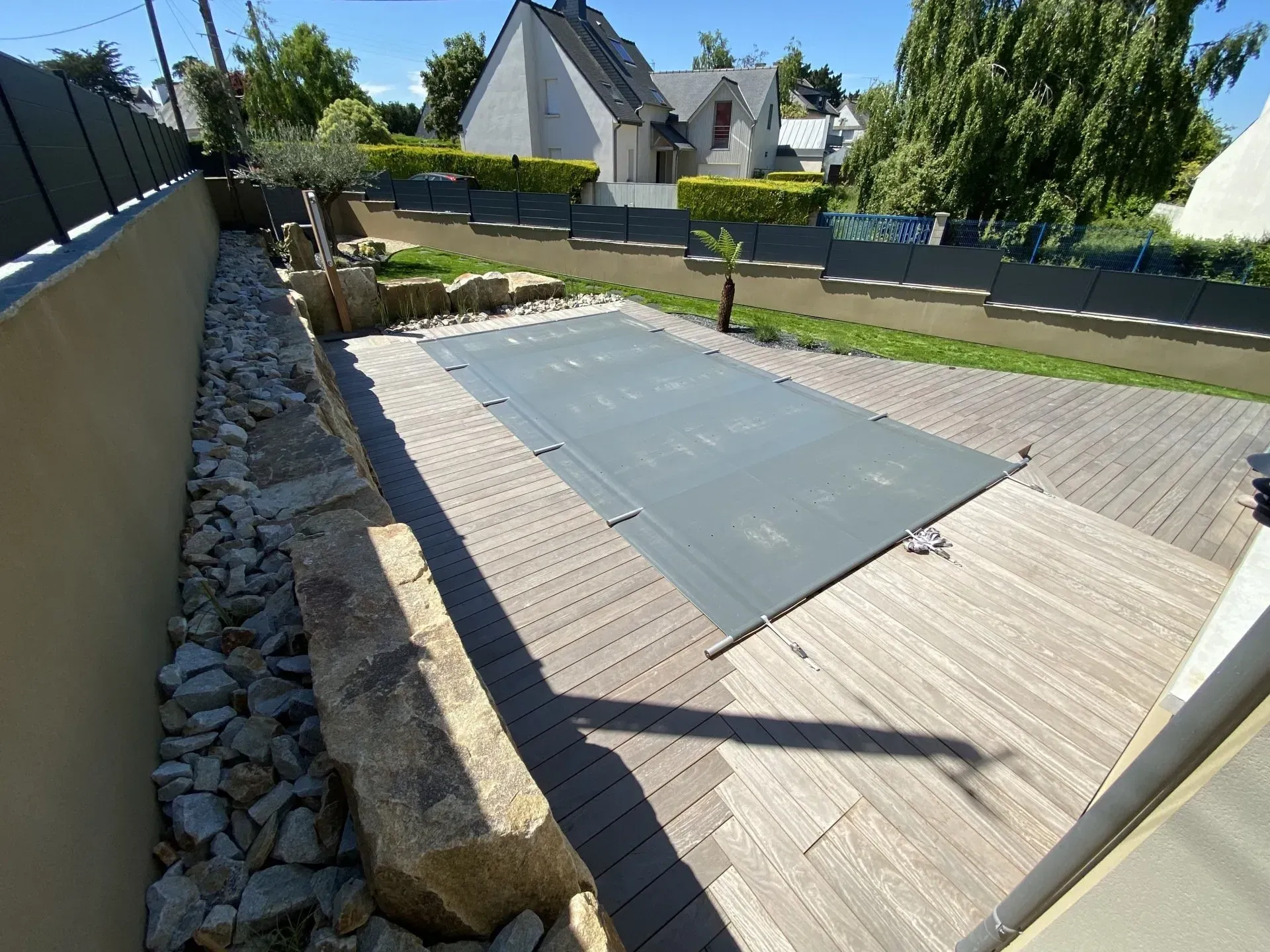 Terrasse arrière avec piscine rectangulaire couverte ou Jacuzzi, bordure en pierre et maisons voisines baignées de soleil.