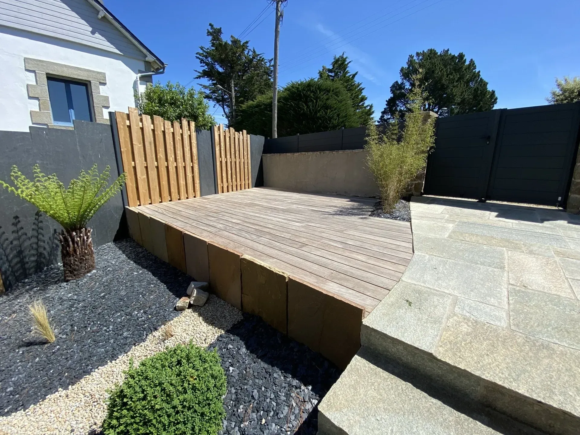 Terrasse en bois surélevée jouxtant un jardin de gravier, avec un portail noir et une rampe en béton dans une cour ensoleillée.