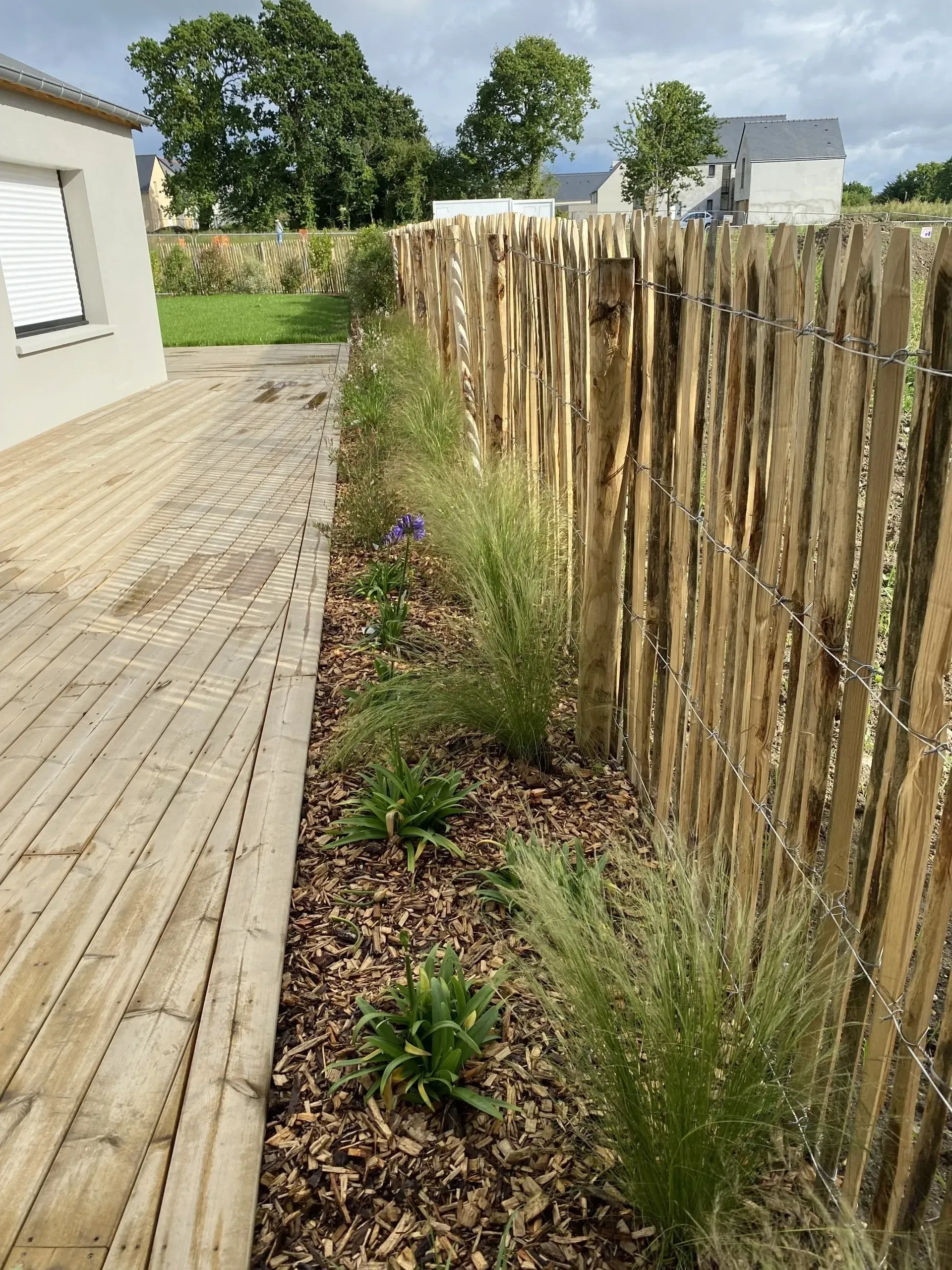 Terrasse en bois jouxtant un parterre de jardin clôturé, orné d'arbustes et de fleurs violettes, par une journée ensoleillée.