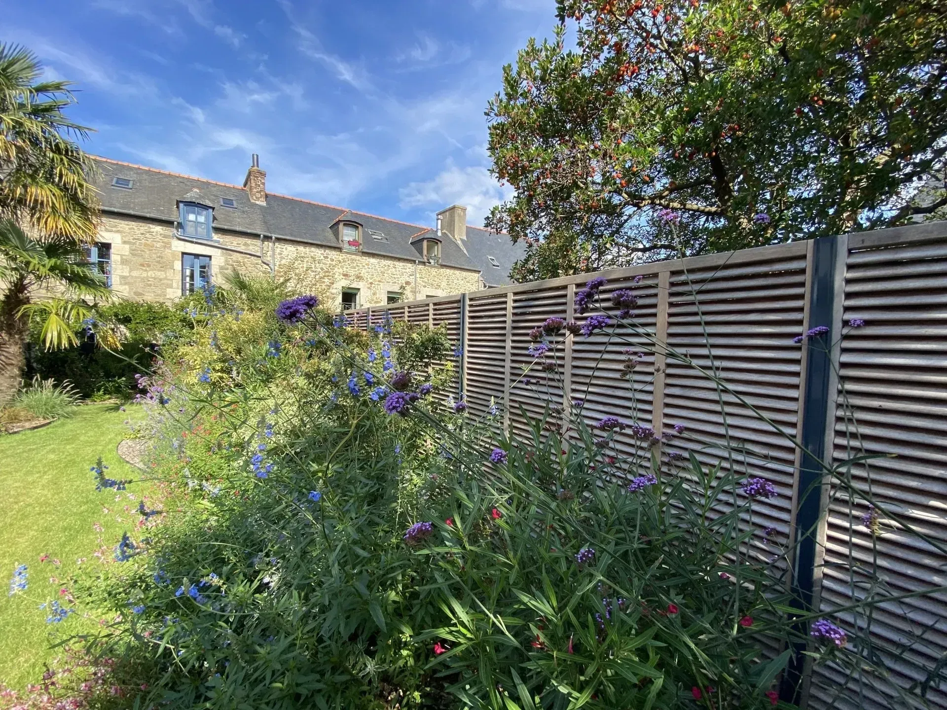 Allée de jardin longeant une clôture en bois et des maisons en pierre sous un ciel bleu.