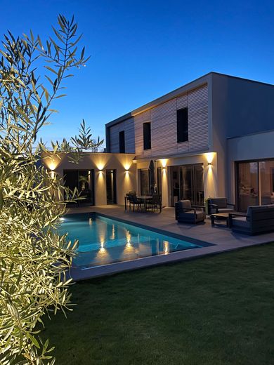 Maison moderne avec piscine et terrasse éclairées au crépuscule, encadrées par un arbre et une pelouse.