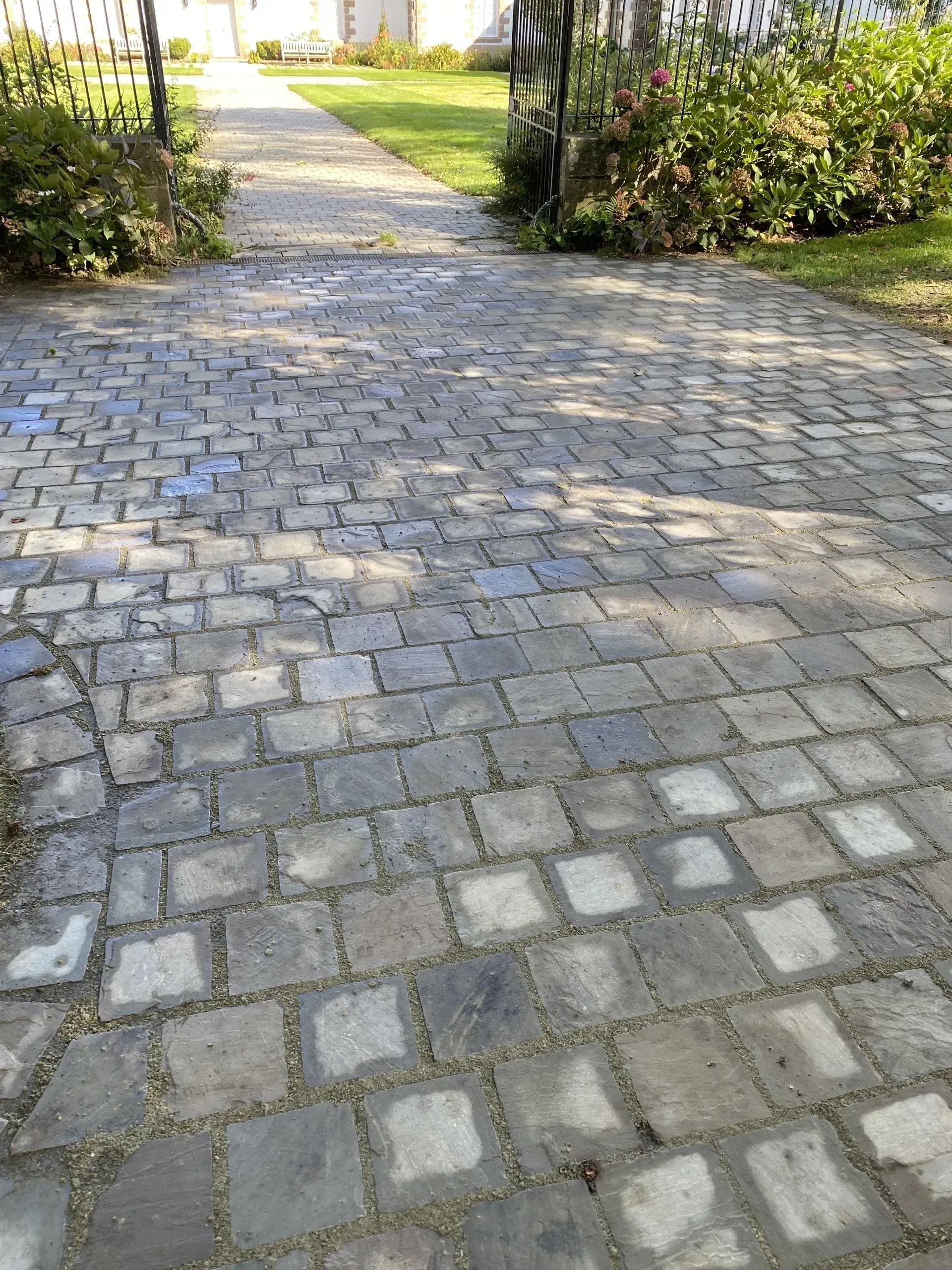 Allée de jardin pavée de pierres, bordée de haies et de verdure de part et d'autre.