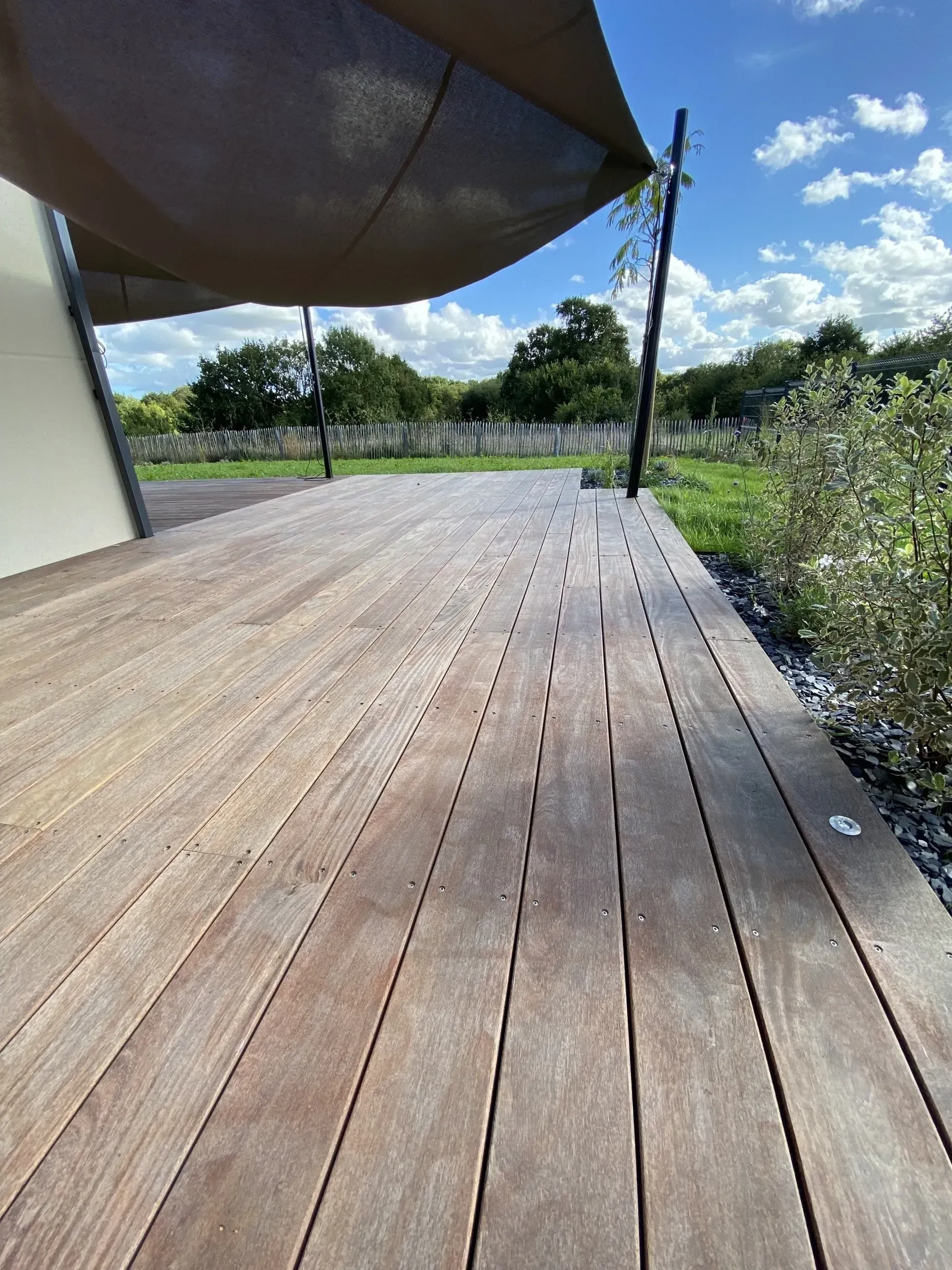 Terrasse en bois le long d'un mur blanc, ombragée par un auvent, avec de la verdure et un ciel ouvert au-delà.