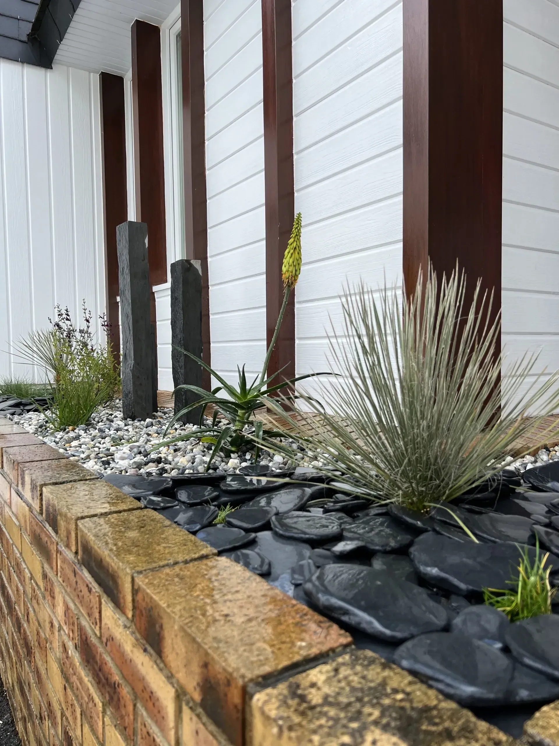 Jardin paysager moderne avec roches sombres, herbes et poteaux en bois le long d'un mur blanc.