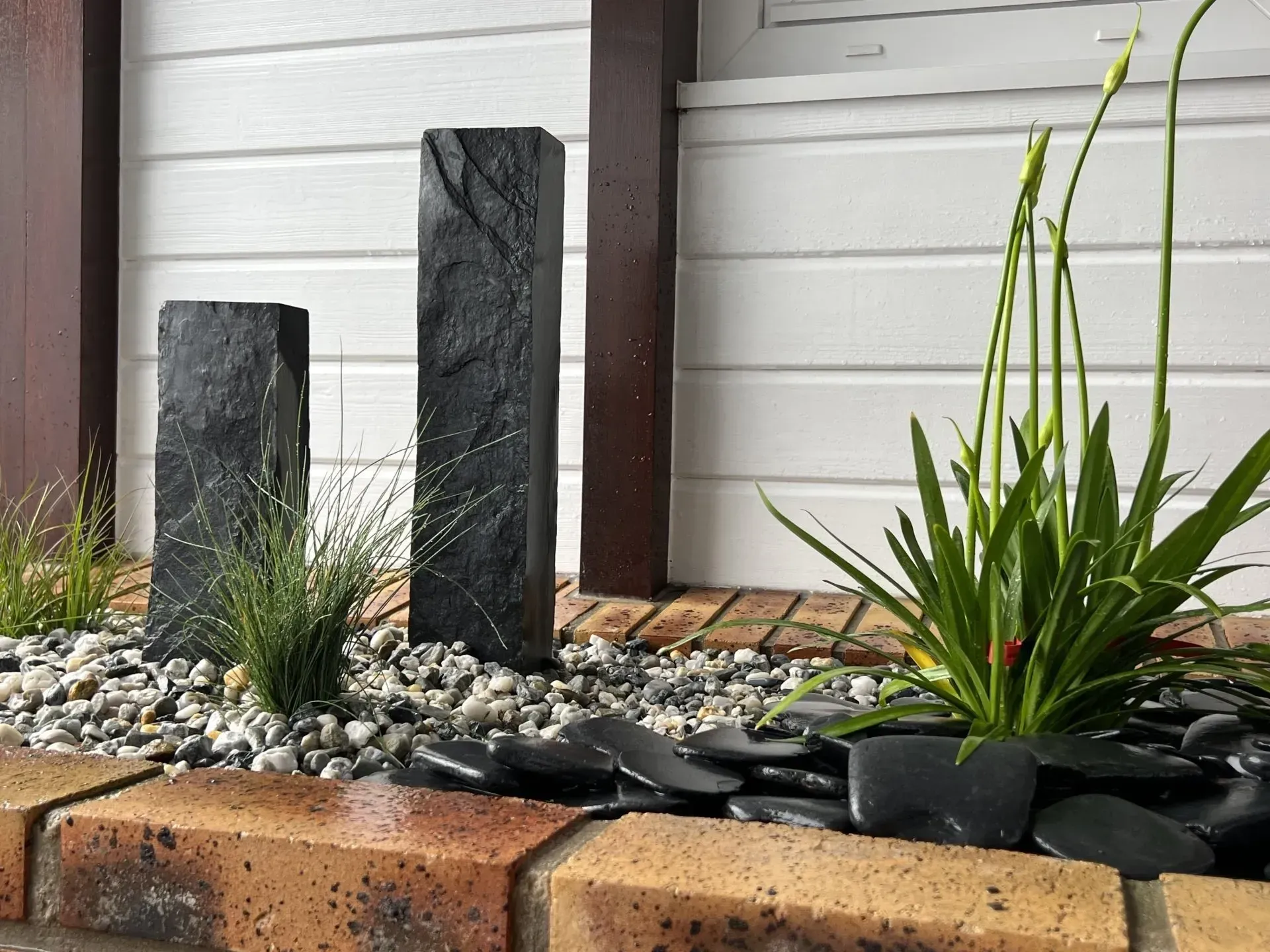 Jardin de rocaille moderne en façade, avec colonnes en pierre noire et plantes vertes en pot sur fond de bardage blanc.
